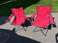 Two red folding camping chairs side by side on pavement with grass in background. Both have fabric seats, cup holders on right armrests, and metal folding legs.