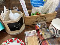 Wooden utensil holder with labeled compartments containing white plastic spoons and forks, with packaged bamboo skewers and colorful straws beside it.