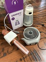Zappify Z20 mosquito zapper with box, small grey portable fan, rose gold and white karaoke microphone with USB cable, displayed on wooden surface