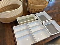 Photo showing two baskets and five white plastic drawer organizers arranged on a wooden surface.