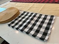 Two table runners, one red and black buffalo plaid and one beige, displayed flat on a surface with black and white buffalo plaid placemats and round natural woven placemats stacked at the edge.