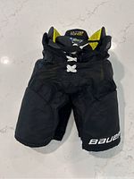 Front view of black Bauer youth hockey pants with visible lace-up front, yellow interior padding slightly visible, and Bauer logo on lower leg.