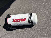 Side view of the white RDX youth punching bag laying on pavement with visible black, red markings and carry handle.