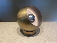 Front angled view showing the brass spherical orb shape with concave reflector and central chrome orb element, sitting on a cylindrical brass base.