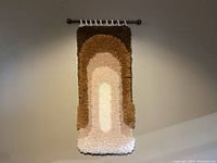 Full view of the hammock style latch hook tapestry with wooden dowel on wall. Displays geometric arch design in earth tones.