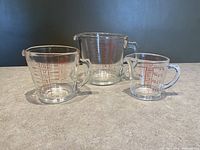Set of three clear glass Pyrex measuring cups placed side by side showing graduated red markings and handles