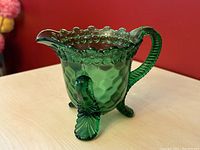 Front angle of vintage green glass creamer dish showing textured body, floral rim, three leaf feet, and handle.