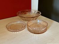 Three vintage pink Depression glass dishes displayed on wood table: floral bowl, sawtooth edged small plate, covered sawtooth dish with lid