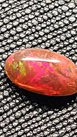 Close up photo of 1.12ct oval cabochon chocolate opal showing bright red and green play of color on black textured background.