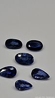 Five natural Ceylon blue sapphires displayed on a grey surface. Mixed shapes including oval and pear cut stones with dark to medium blue color.