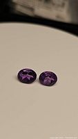 Pair of oval purple amethyst gemstones placed on a neutral beige background, showing the deep violet color and faceted oval cut shape.