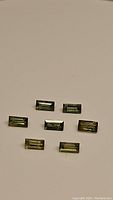 Seven natural green baguette cut sapphires laid out on a beige surface, showing their size, cut, and green color with yellow undertones.