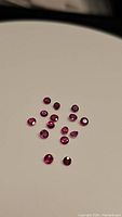 Photo showing 14 small natural rubies with deep red, pink, and purple colors, round and faceted, approximately 2mm each.