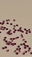 Close-up photo showing many small, oval and round natural pink sapphires scattered on a beige background.