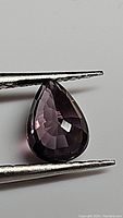 Close-up photo of the natural purple spinel pear cut gemstone held by tweezers, showing the faceted surface and deep purple color.