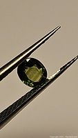 Close-up image of a faceted oval-shaped green-yellow sapphire gemstone held by tweezers.