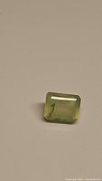 Close-up image of a mossy green rectangular emerald cut rutilated prehnite gemstone on a neutral background.