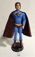 Full body front view of Superman Ken Doll in blue costume with cape standing on a black stand.