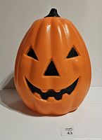 Front view of the orange blow mold Jack O Lantern with carved eyes, nose, and mouth, showing the black stem on top and a measured size card nearby.