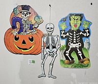 Three vintage Halloween diecut decorations hanging on a wall for display: skeleton in pumpkin, full skeleton, and Frankenstein skeleton.