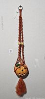 Macrame holder with ceramic pumpkin planter suspended inside, showing hanging loop and tassel detail.