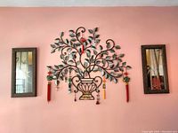 Front view of the decorative iron wall art piece in center with floral design and hanging tassels flanked by two framed prints on a beige wall.