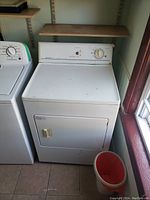 Front and partial side view of white Frigidaire Crown heavy duty electric dryer. Control panel with dial and indicator light visible. Dryer door closed with handle on front.
