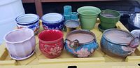 Various pots and planters shown on yellow tray including red ceramic, blue and white ceramic, blue ceramic with flower design, brown glazed, green glazed plastic, pink glazed with ridges, and small flower-shaped planters.