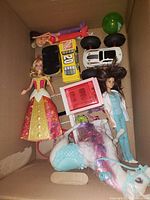 Photo showing a box with various toys: dolls, toy vehicles, a pink toy oven, and a multi-colored plush toy.