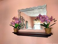 Photo showing Venetian style mirror with scalloped edges and antique phoenix design above dark wooden mantle shelf, flanked by two artificial orchid plants with purple flowers in brown pots.