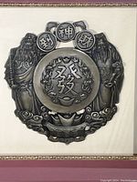 Close-up of metal medallion showing two robed figures, Chinese characters in center and on circular medallion tops, set on textured beige background.
