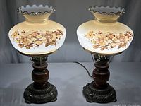 Pair of vintage parlor lamps with ruffled top cream glass globes decorated with floral designs, wooden bases and brass ornate footings, set side by side for scale and condition