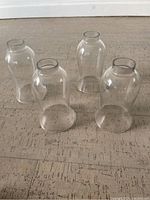 Four clear glass hurricane lamp shades displayed upright on a textured surface showing overall shape and size.