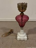 Full view of cranberry glass table lamp with white base and brass metal holder on top, placed on floor.