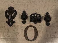 Five iron pieces on a wooden surface: three ornate cast iron decorative hardware pieces, one iron number zero, and one cast iron piece with a pattern possibly a trivet.