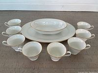 Full view of 8 white Lenox Oxford bone china teacups arranged around a large oval serving plate and a serving bowl, all with gold trim.