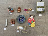 Overview of all items laid on table showing bird cup, wooden mask, cloisonné plate, wooden shoes, fabric doll, jewelry box with earrings, tassels, knife, and container.