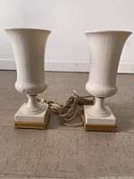 Front view of the pair of white Lenox table lamps showing fluted urn-shaped bodies and gold-accented square bases with attached cords.