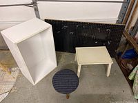 Collection of four items on concrete garage floor: white cabinet, black pegboard, small off-white foot stool, round patterned upholstered foot stool.