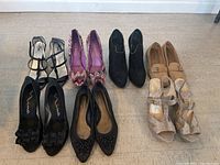 Six pairs of women's shoes arranged in two rows on floor: black T-strap heels, multicolor pumps, black suede ankle boots, tan flats, and two pairs of black flats with differing decorations.
