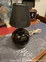 Black round boudoir table lamp with shiny spherical base and black fabric empire shade with small dent, placed on patterned grey cloth and wooden surface