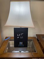 Brass colored table lamp on glass table with a white fabric shade, showing the whole lamp including the base and shade.