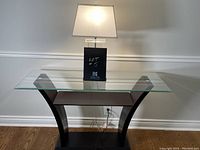 Console table showing clear glass top, curved dark wood vertical supports, middle horizontal shelf, and platform base on wood floor against wall.