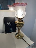 Full view of vintage faux lantern table lamp lit, showing frosted glass shade with pink ruffled edge and embossed design, brass-tone base with knob, electrical cord visible.