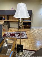 Full view of the brass-colored floor lamp with white shade, showing lamp in off state and wear on base.