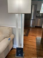 Floor lamp standing next to beige couch on hardwood floor in kitchen/living room setting, showing chrome base and white rectangular pole with lampshade.