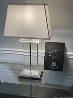 Front view of the glass table lamp powered on showing the rectangular white fabric shade and clear glass column base with chrome accents.