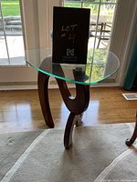 Side coffee table with clear thick glass top and dark wooden curved legs from front angle, showing the sculptural base design
