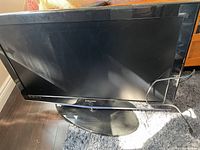Front angle view of black Samsung television on an oval swivel stand, placed on carpeted floor.