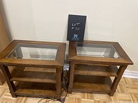 Pair of wood side tables with glass inset tops, placed against wall on parquet floor, showing overall view of both tables side by side.